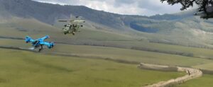 Zwei RC-Hubschrauber fliegen über ein Feld mit Bergen im Hintergrund.